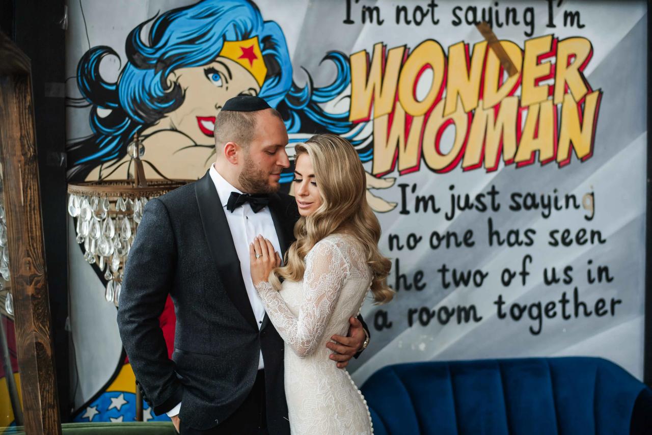 Frum wedding couple sharing an affectionate embrace in front of a “Wonder Woman” themed backdrop.