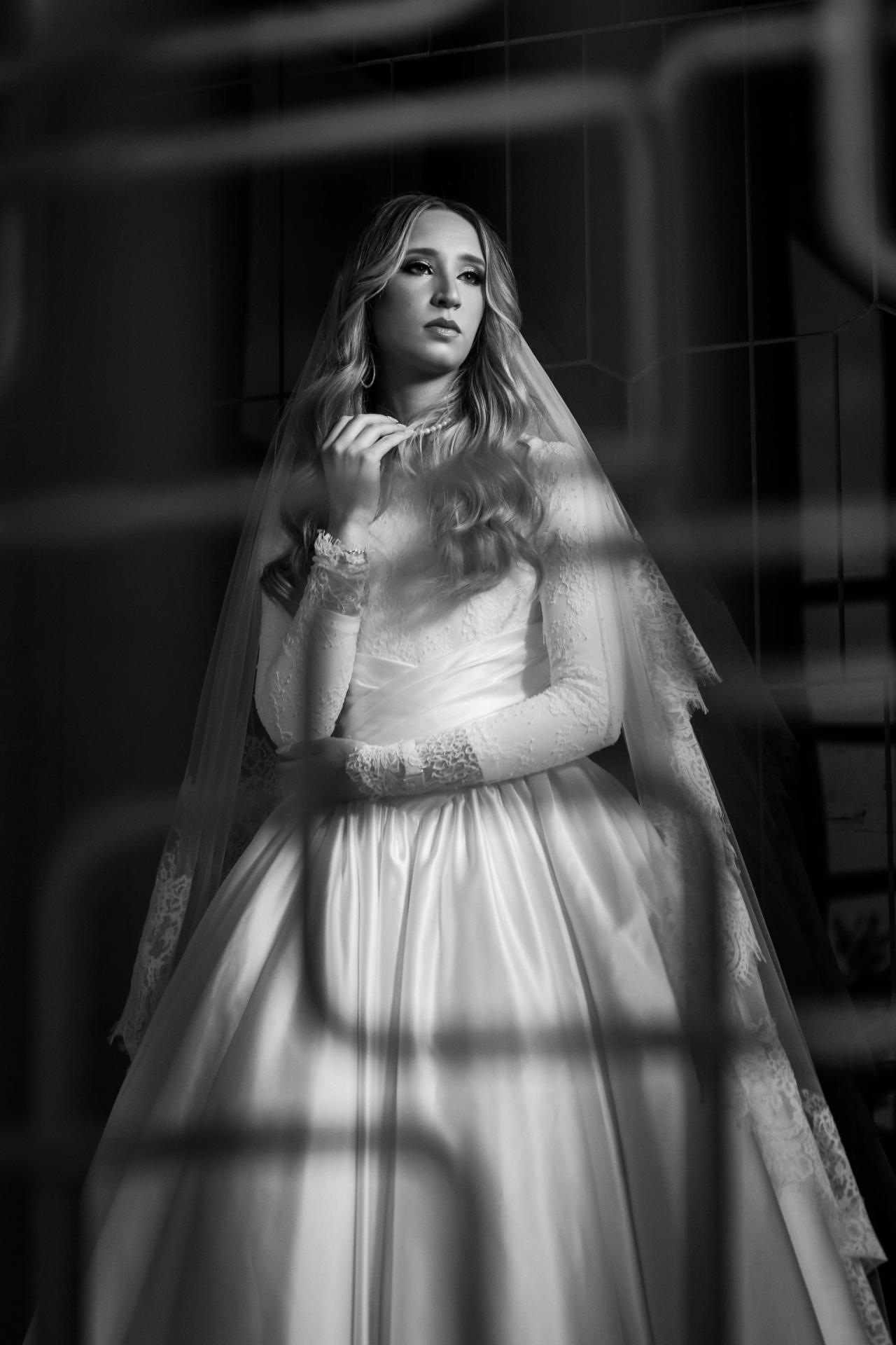 Black-and-white portrait of a bride in a lace dress and veil, holding her bridal shawl and looking calmly at the camera in a dramatic indoor setting.