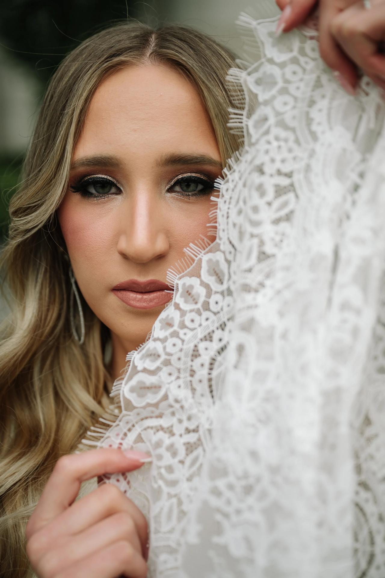Close-up portrait of a bride holding her white lace veil near her face while looking directly at the camera.