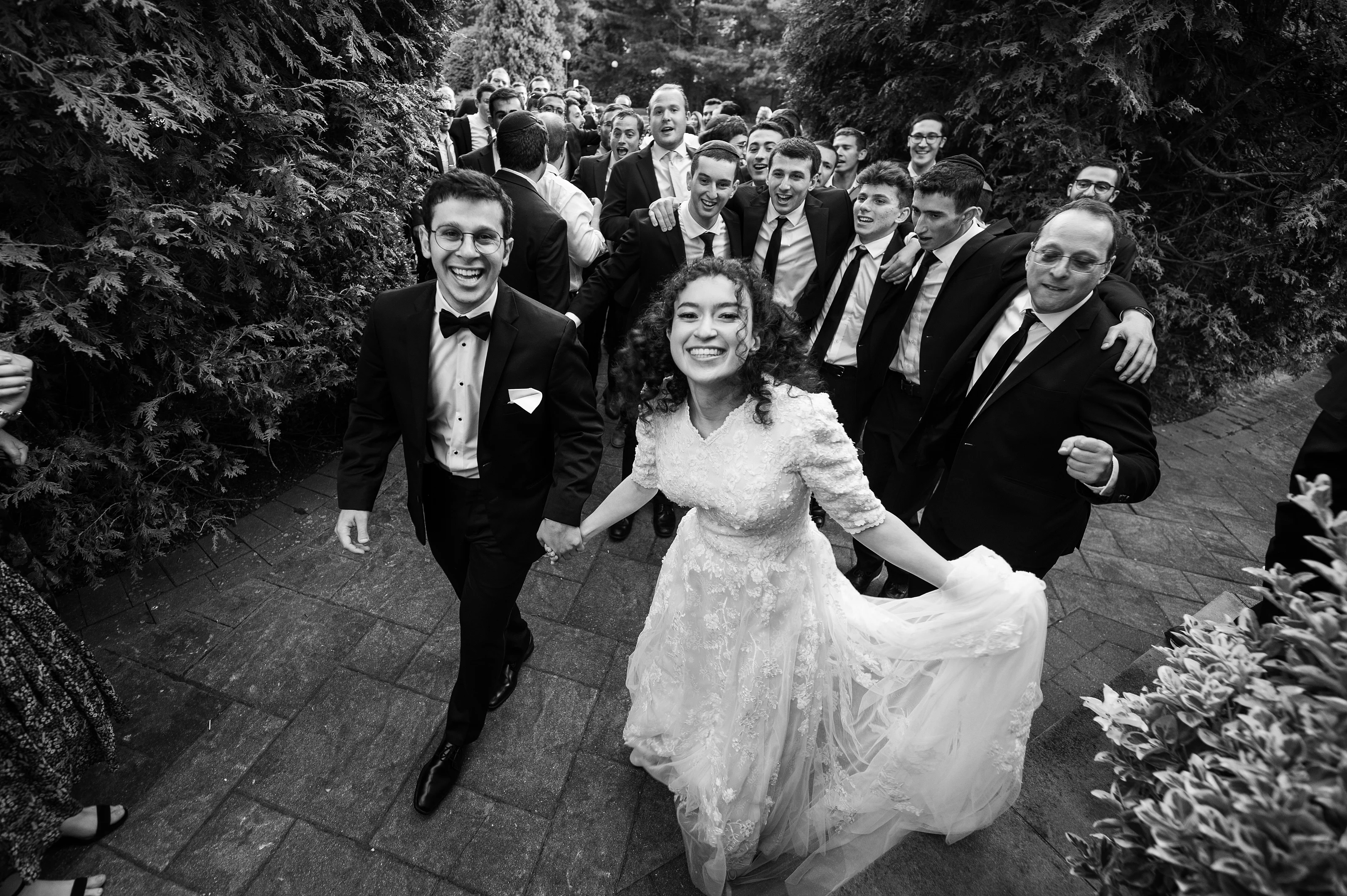 Black-and-white photo of a smiling bride in a lace dress dancing with the groom at an outdoor wedding reception, surrounded by friends in suits.