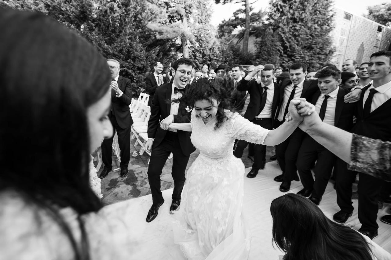 Black-and-white wedding scene outdoors where the bride laughs mid-dance while wedding guests in suits form a cheering circle around her.