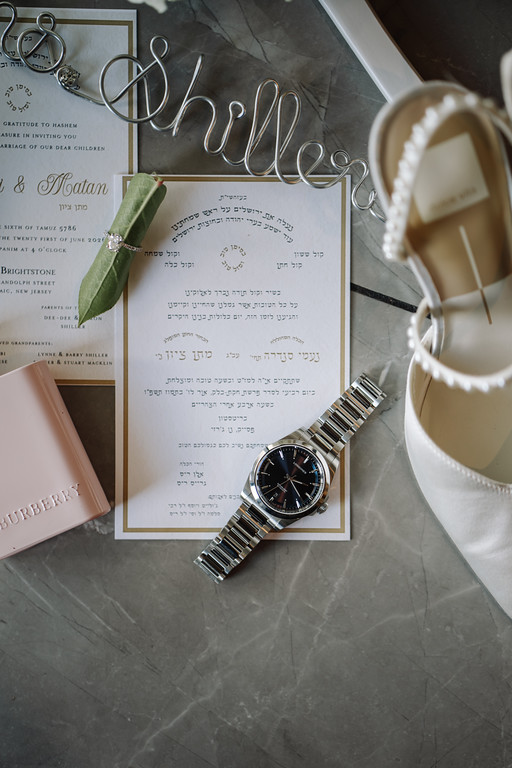 Flat-lay detail of a ketubah page surrounded by a watch, jewelry, and the bride’s white heels on a textured gray surface.