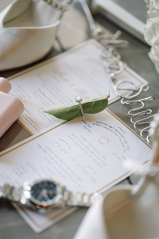Close-up of event invitation papers with a small greenery sprig pinned on top and wedding accessories softly blurred in the background.