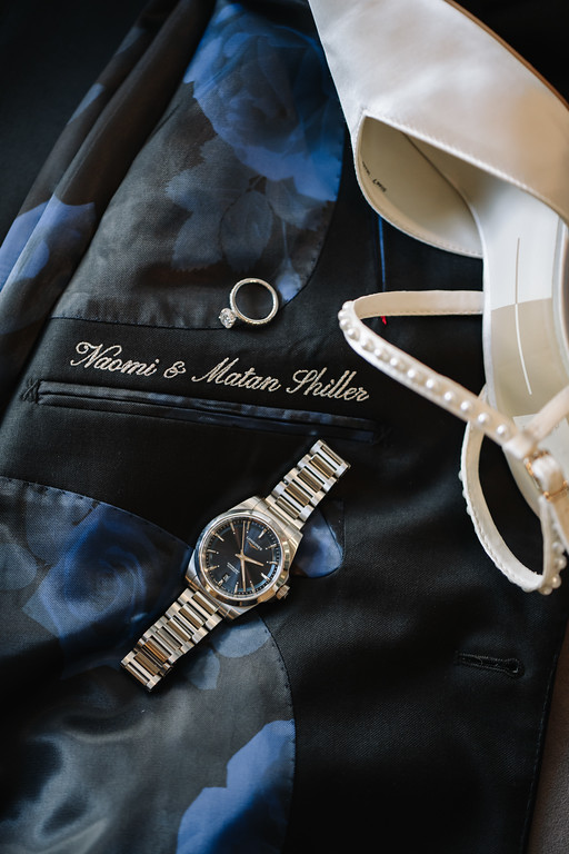 Close-up detail of a suit, including a wristwatch, a ring, and a decorative shoe, arranged for a wedding day flat lay.