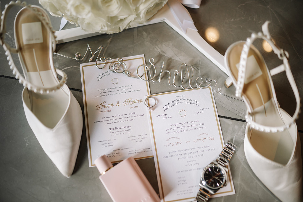Close-up of a wedding invitation card titled “Matan & Matian,” surrounded by bridal heels, jewelry, and a watch on a gray stone surface.