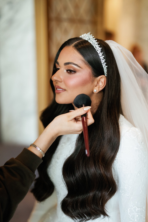 Bride in a side profile wearing a jeweled headband and veil as a makeup artist applies makeup with a brush.