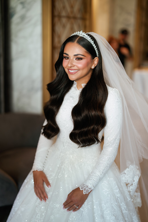 Bride in a white lace outfit and veil, smiling at the camera indoors with soft background blur.