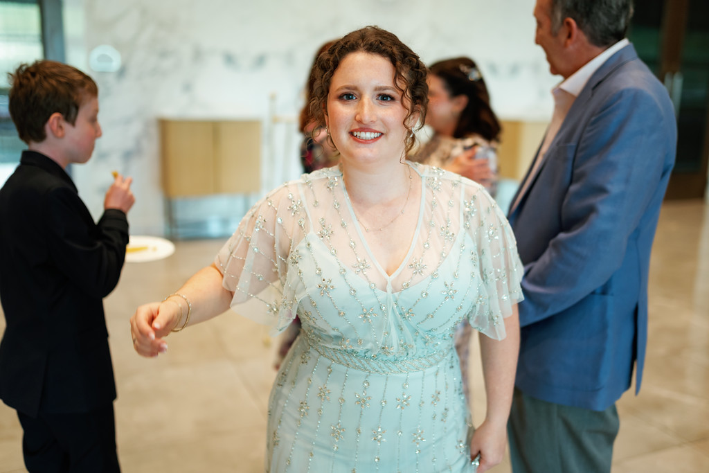 Bride in a light blue embellished dress smiling while dancing at a celebration, with guests in the background clapping and talking.