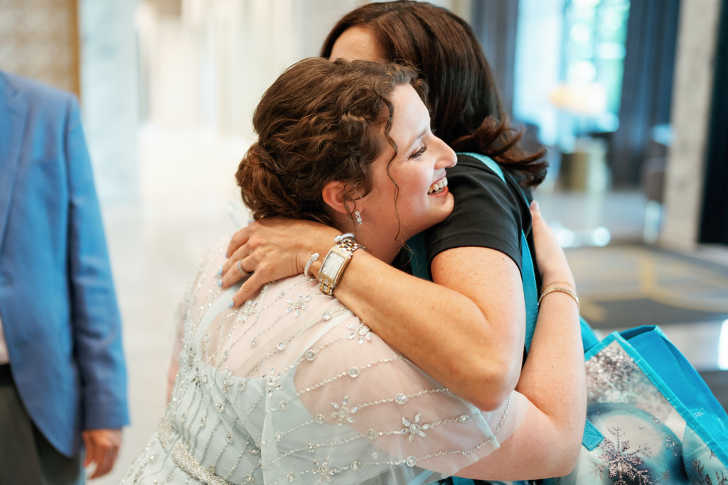 Two people share a close hug during a wedding event, smiling as they embrace in a softly lit indoor setting.
