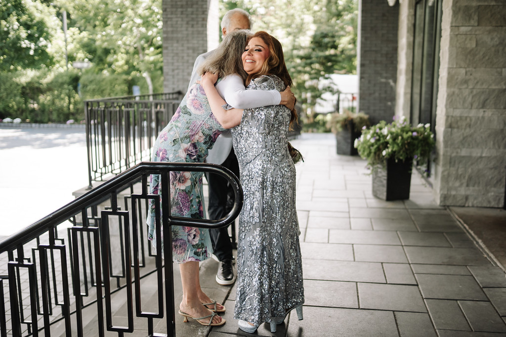 Bride in a silver sequined dress embraces a seated relative in the outdoor courtyard, with a joyful smile and arms wrapped around her.