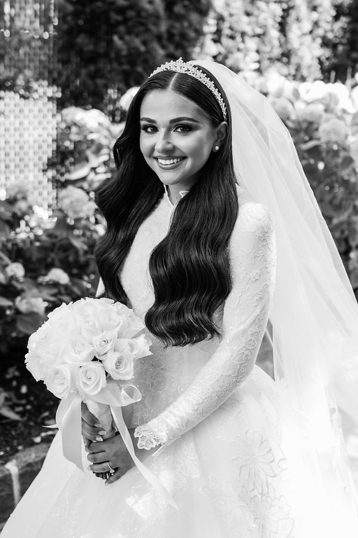 Black-and-white portrait of a smiling bride outdoors, wearing a veil and holding a bouquet of white roses.