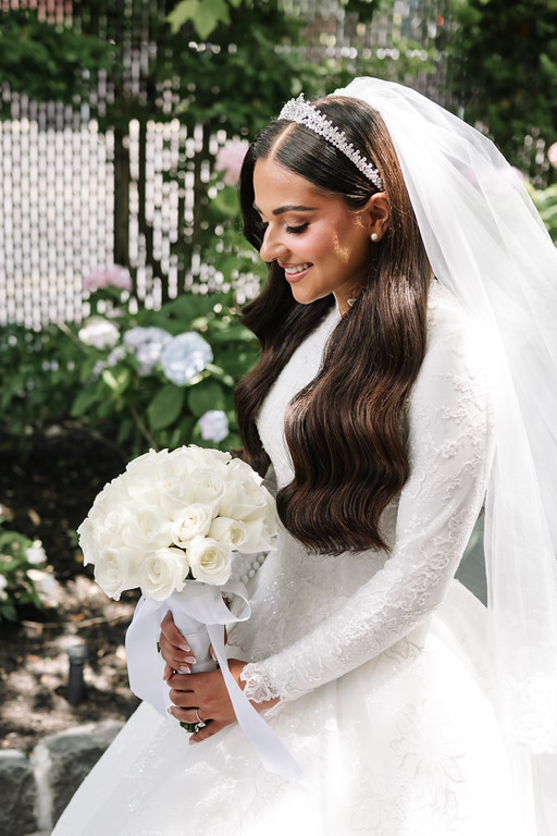 Bride in a white veil and lace dress smiling while holding a bouquet of white roses in a garden.