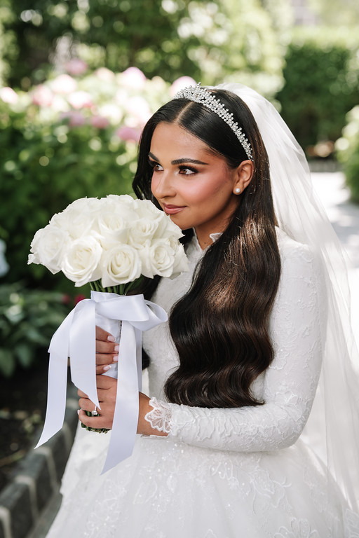 Outdoor wedding portrait of a bride in a white veil and lace gown holding a bouquet of white roses, looking over her shoulder with soft greenery in the background.