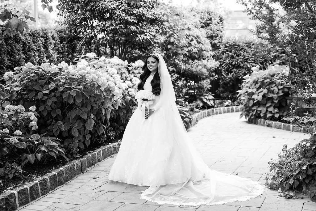 Black-and-white portrait of a bride wearing a flowing gown and veil, standing on a garden pathway with lush flowers and greenery on both sides.