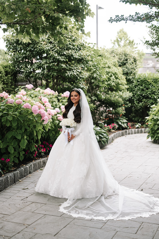 Bride in a white wedding gown and veil holding a bouquet, standing on a garden walkway with pink and white hydrangeas and greenery in the background.