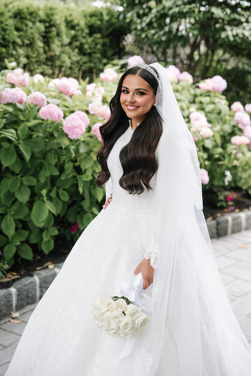 Bride in a white veil and lace gown smiling outdoors in a garden of pink and white flowers while holding a bouquet.