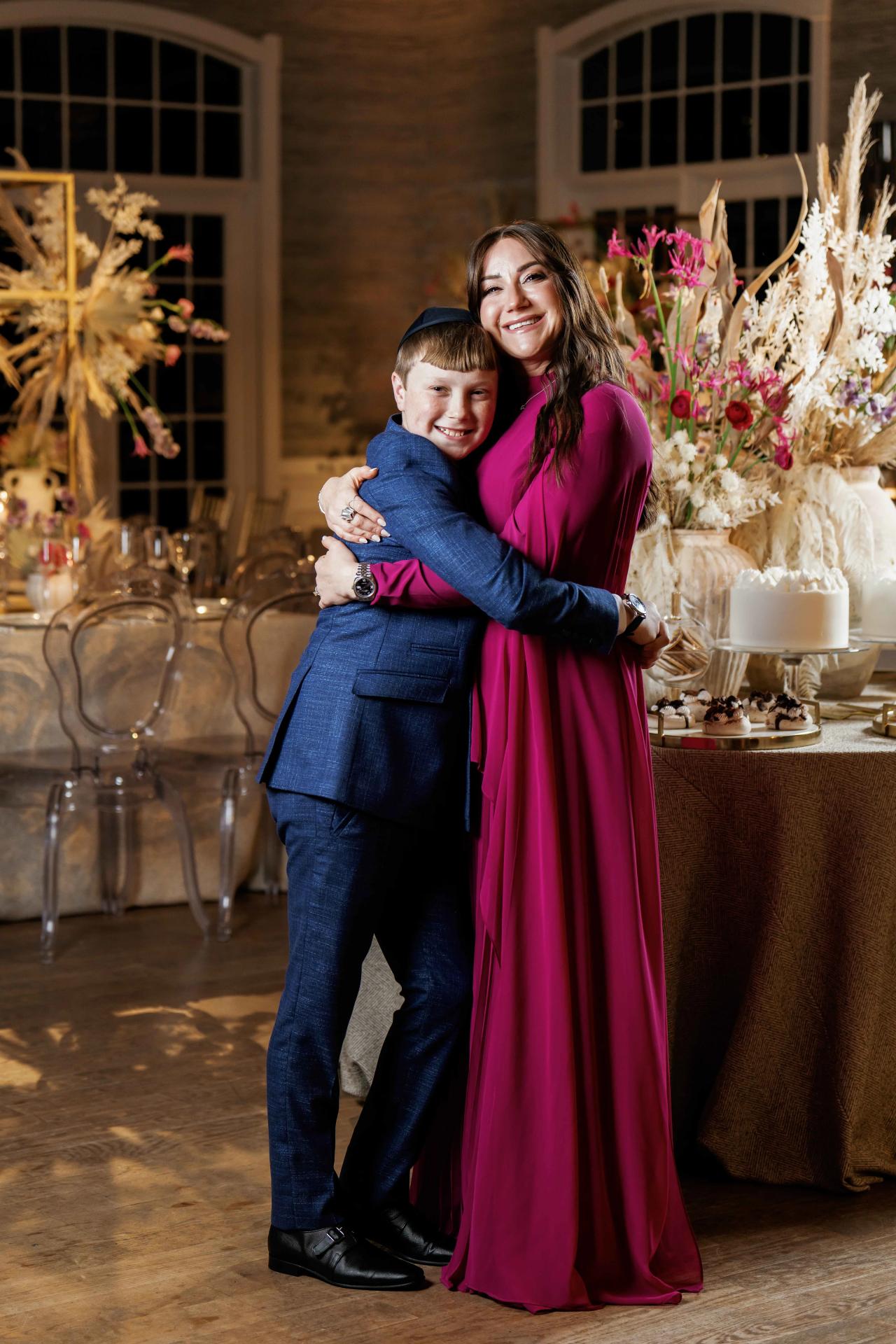 Smiling woman in a magenta dress embraces a young boy in a blue suit in front of a decorated dessert table at a wedding celebration.