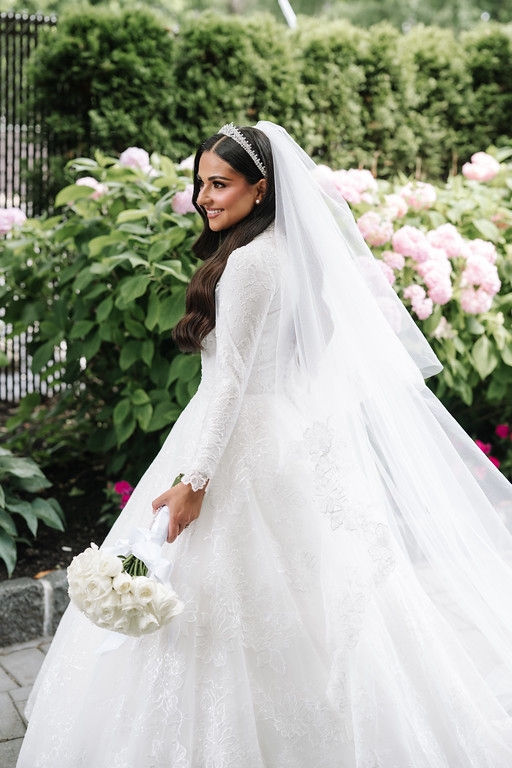 A smiling bride in a white lace gown and veil holding a bouquet on a garden walkway filled with pink and white flowers.