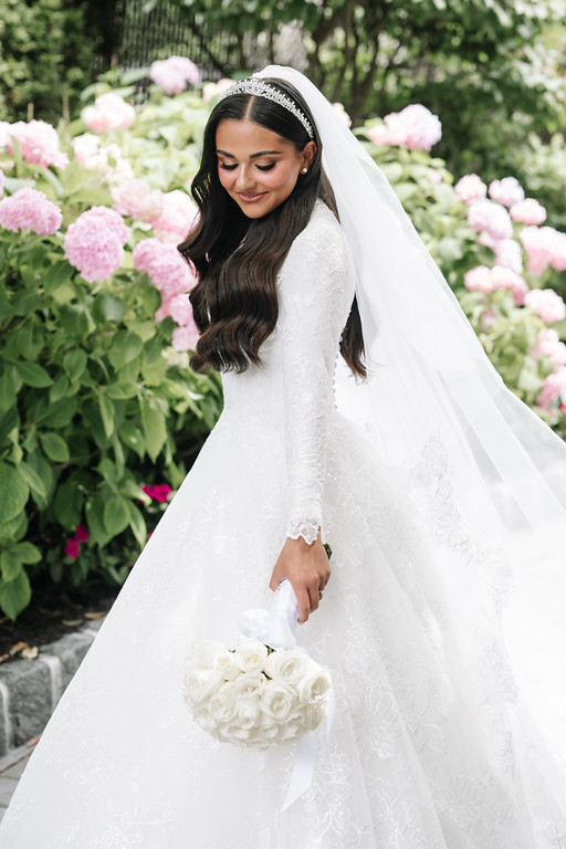 Bride in a white lace gown and veil standing in a garden filled with pink flowers, looking down while holding a bouquet of white roses.