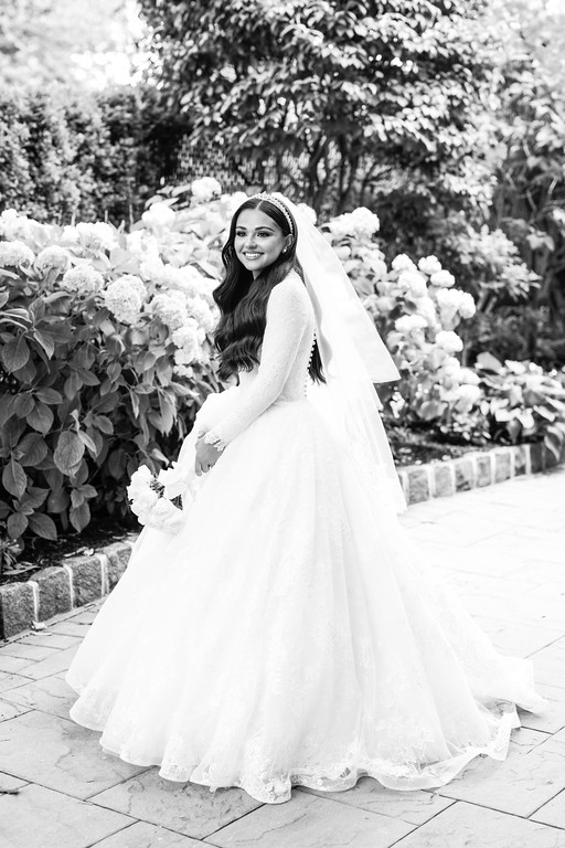 Black-and-white portrait of the bride in a white wedding dress and veil smiling outdoors among abundant white flowers and greenery.