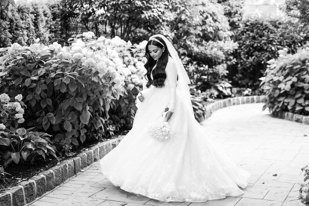 Black-and-white portrait of a bride in a white gown and veil walking along a garden path beside large blooming bushes, looking down as her dress flows.