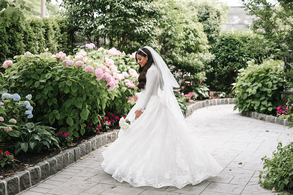 Bride in a white wedding gown and veil walking along a garden path surrounded by blooming hydrangea bushes, looking down softly.