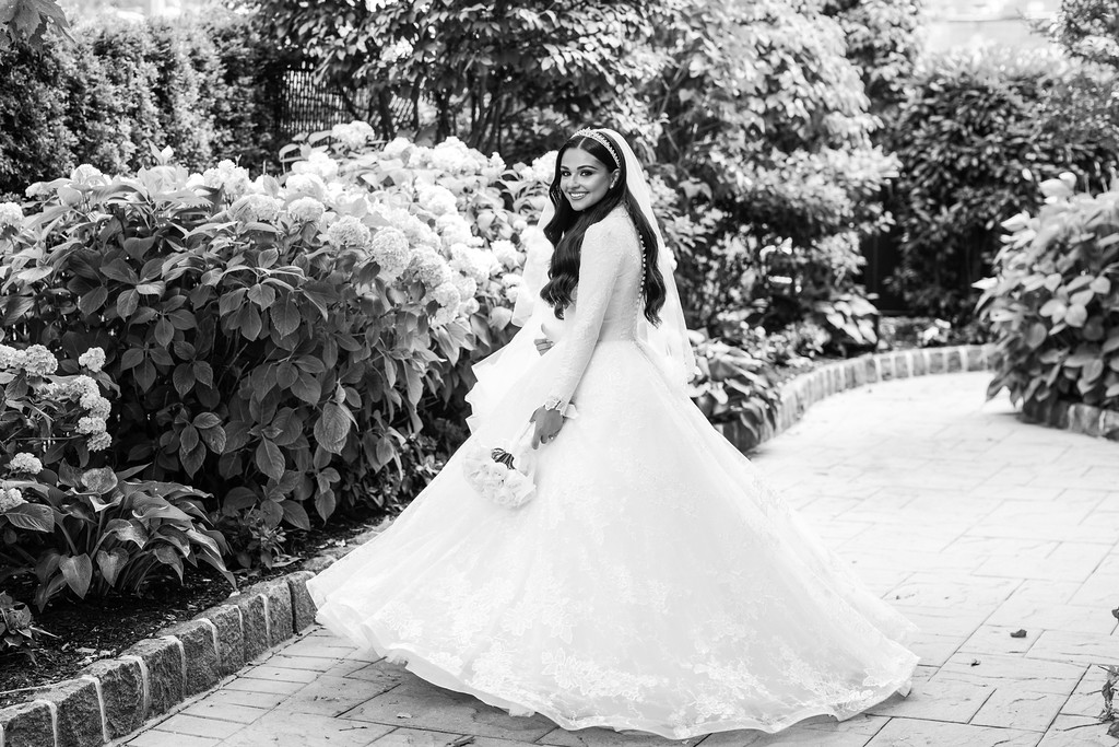 Black-and-white portrait of a smiling bride in a flowing gown standing on a paved garden walkway beside flowering hedges.