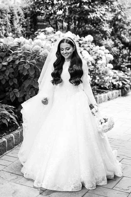 Black-and-white photo of a smiling bride in a lace gown and veil walking along a garden pathway beside flowering hedges.