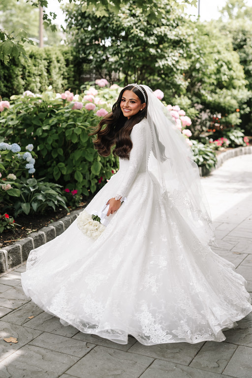 Bride in a white wedding dress and veil smiling while walking along a landscaped garden path with pink flowers in the background.