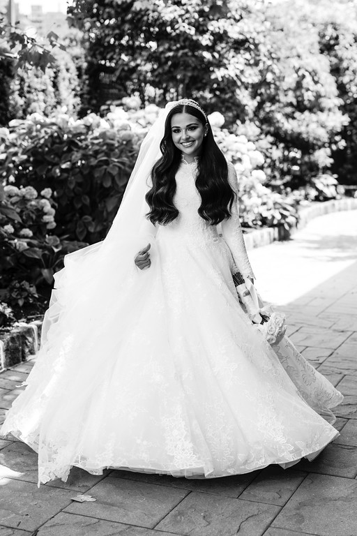 Black-and-white portrait of a smiling bride in a lace gown and veil standing outdoors along a stone walkway beside flowering bushes.