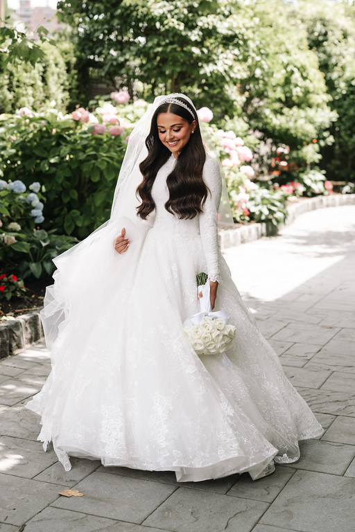 Frum bride in a white lace gown and veil smiling as she walks along a garden pathway with pink flowers behind her, holding a bouquet.
