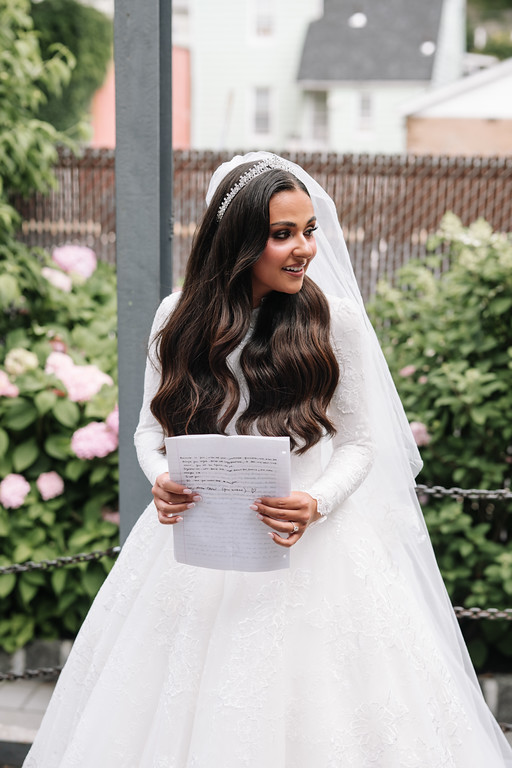 Frum bride in a white lace gown and veil holding a written document at an outdoor garden wedding, smiling softly as she reads.