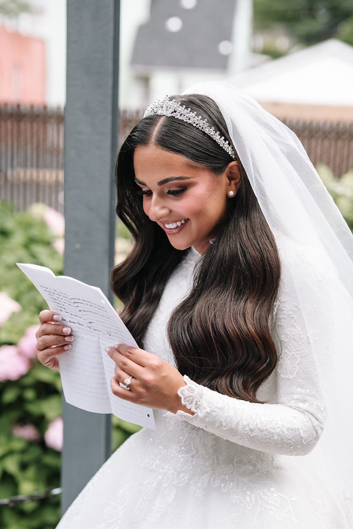 Frum bride in a white gown and veil smiles while reading a paper/ketubah outdoors, holding the pages with both hands.