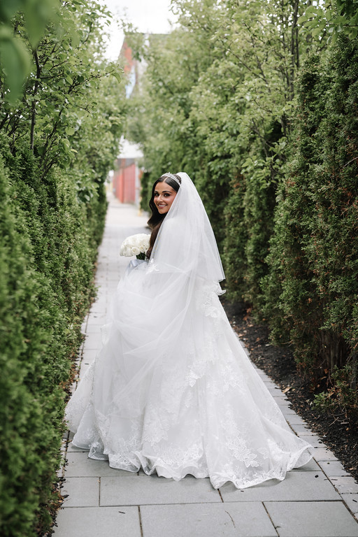 Bride in a white gown and veil walking along a garden walkway, framed by tall green hedges, smiling toward the camera.
