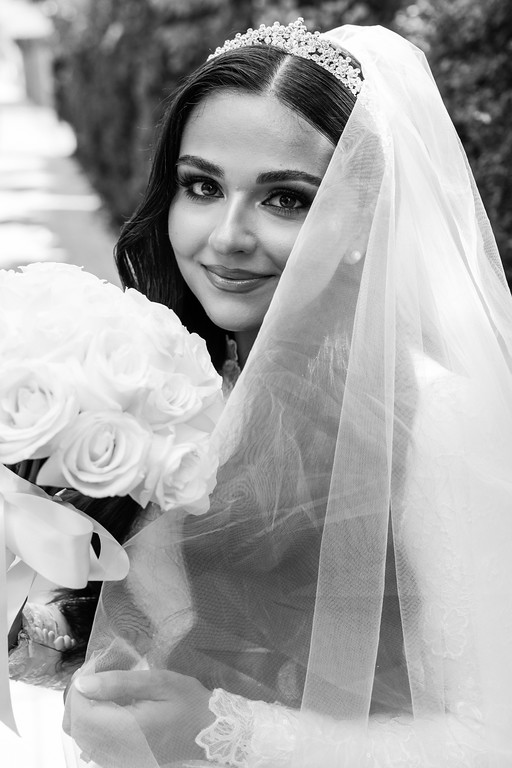Black-and-white portrait of a bride smiling under a lace veil, holding a bouquet with soft floral details in the foreground.