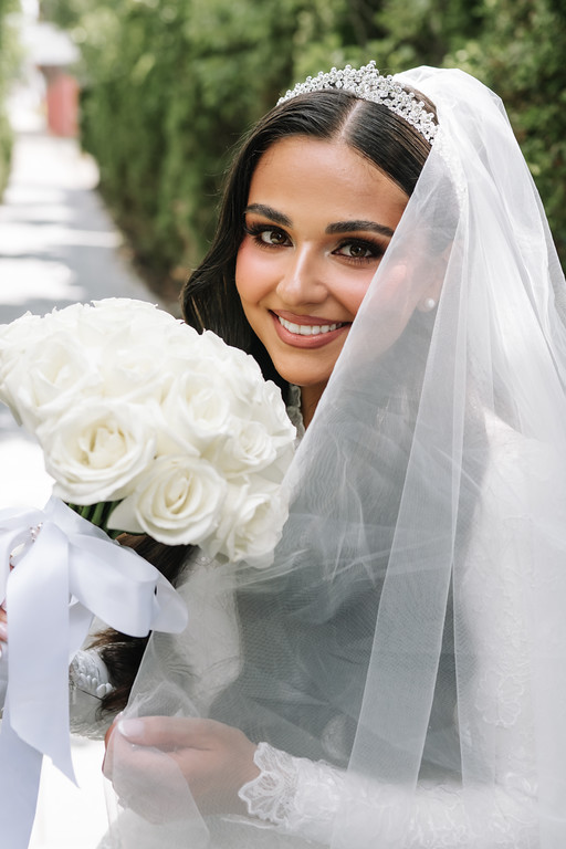 Bride in a white wedding veil smiling toward the camera while holding a bouquet of white roses outdoors.