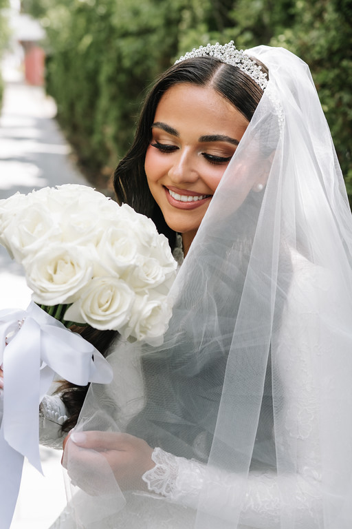 Frum bride in a white headpiece and veil smiling while holding a bouquet of white roses outdoors.