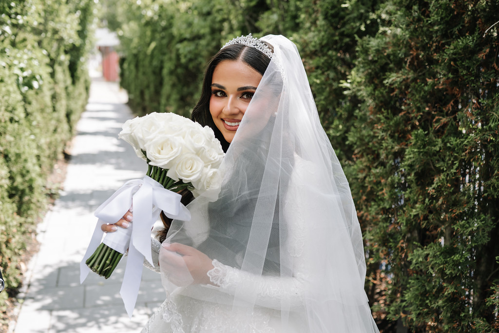 Bride in a white wedding dress and veil smiles outdoors while holding a bouquet of white roses.