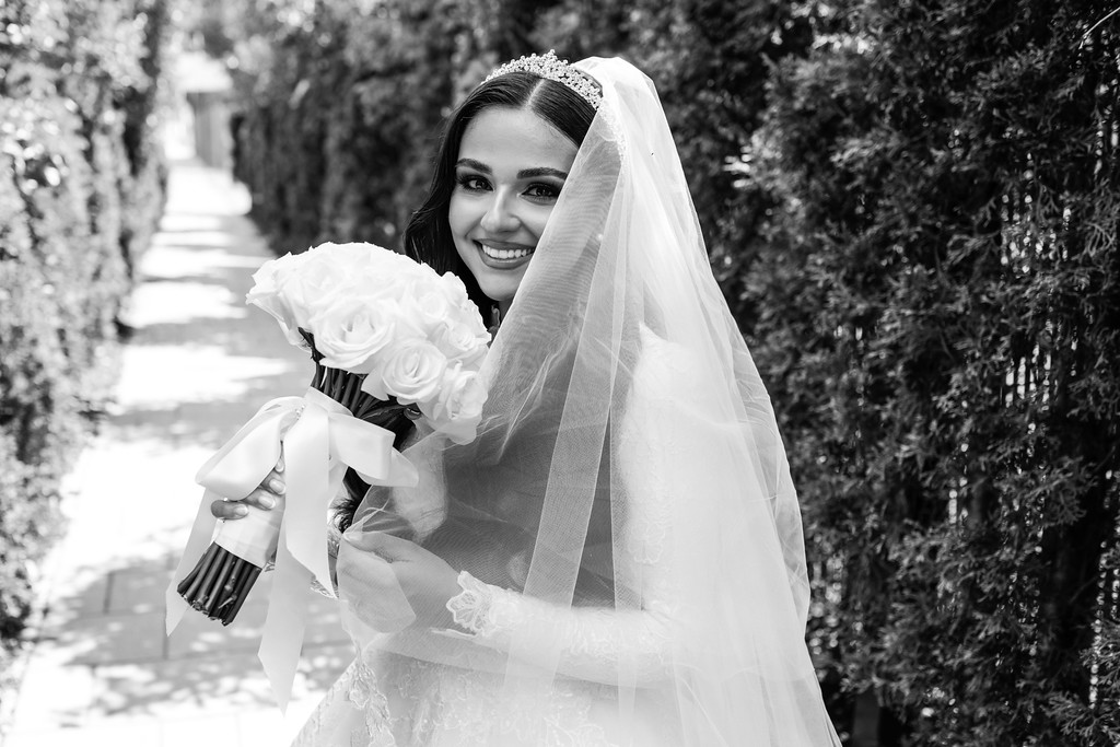 Black-and-white outdoor portrait of a smiling bride wearing a jeweled headpiece and veil, holding a bouquet of white flowers in front of greenery.