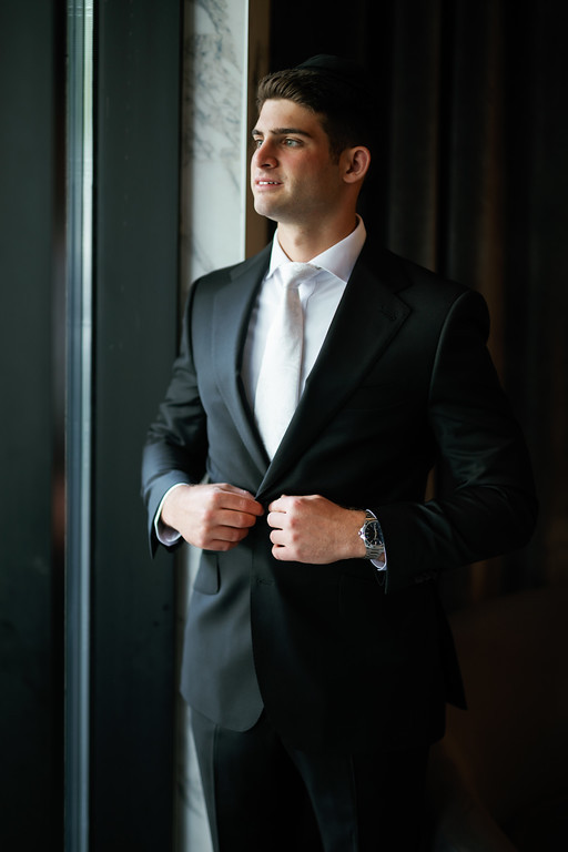 Groom in a dark suit and white shirt smiling while adjusting his jacket near an indoor doorway with soft side light.