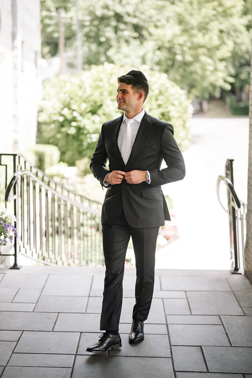 Groom in a dark suit smiling and adjusting his jacket outdoors along a walkway with greenery in the background.