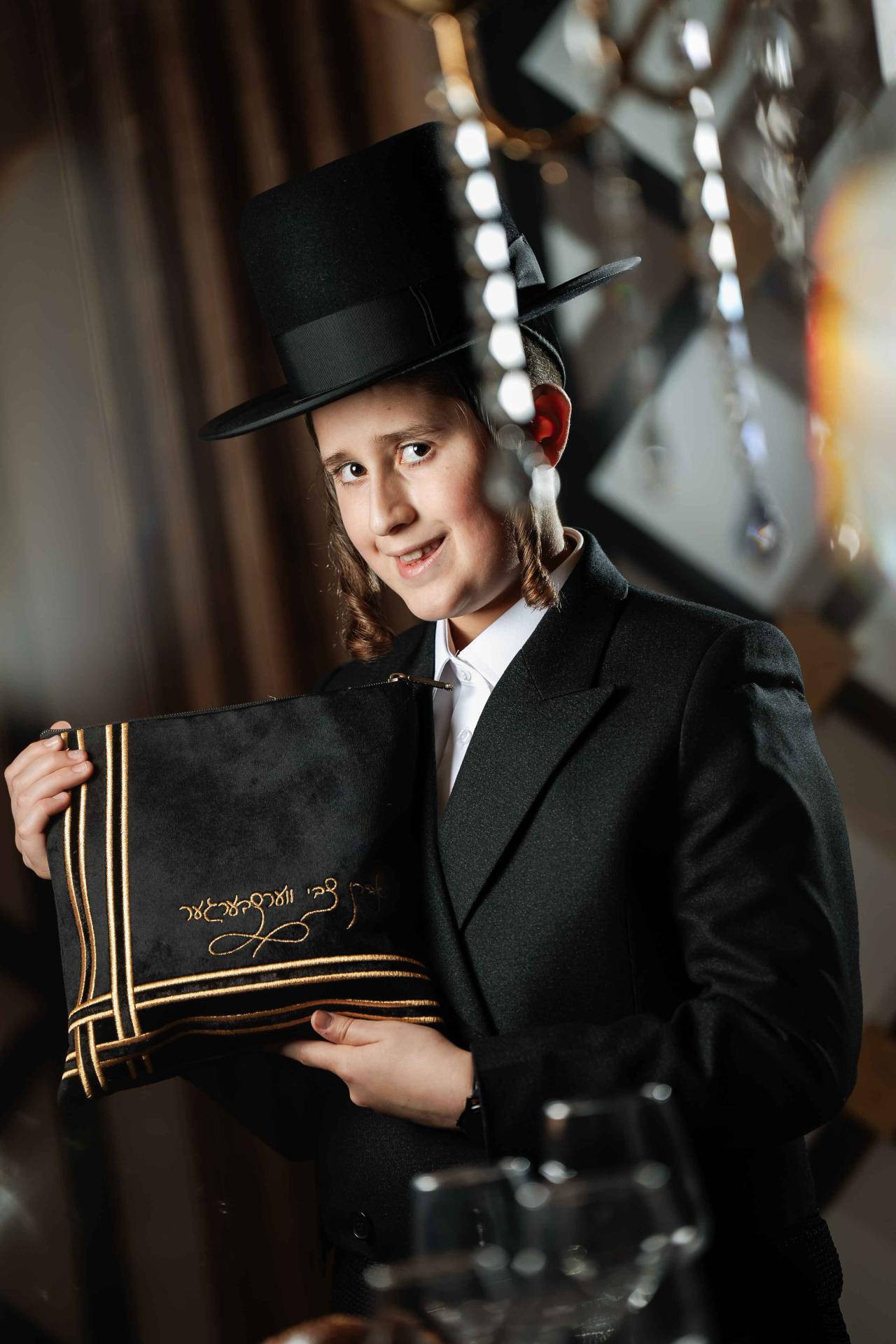 Smiling boy in a black hat and suit holding an embroidered Torah cover in a softly lit synagogue corridor, with decorative bokeh lights in the background.