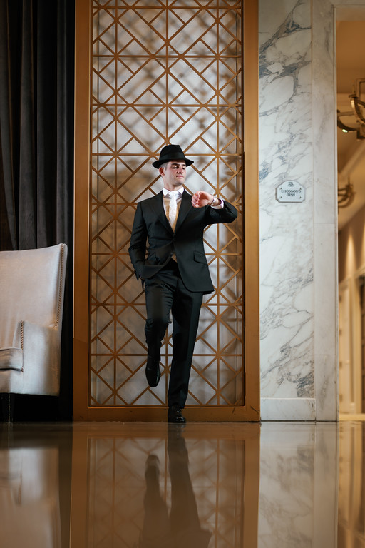 Groom in a black suit and hat posing confidently indoors in front of a decorative gold lattice panel, with the mirrored floor reflecting him.