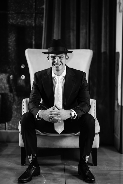 Black-and-white portrait of a groom in a black hat, suit, and tie sitting centered on a cushioned chair, smiling toward the camera in a formal indoor setting.