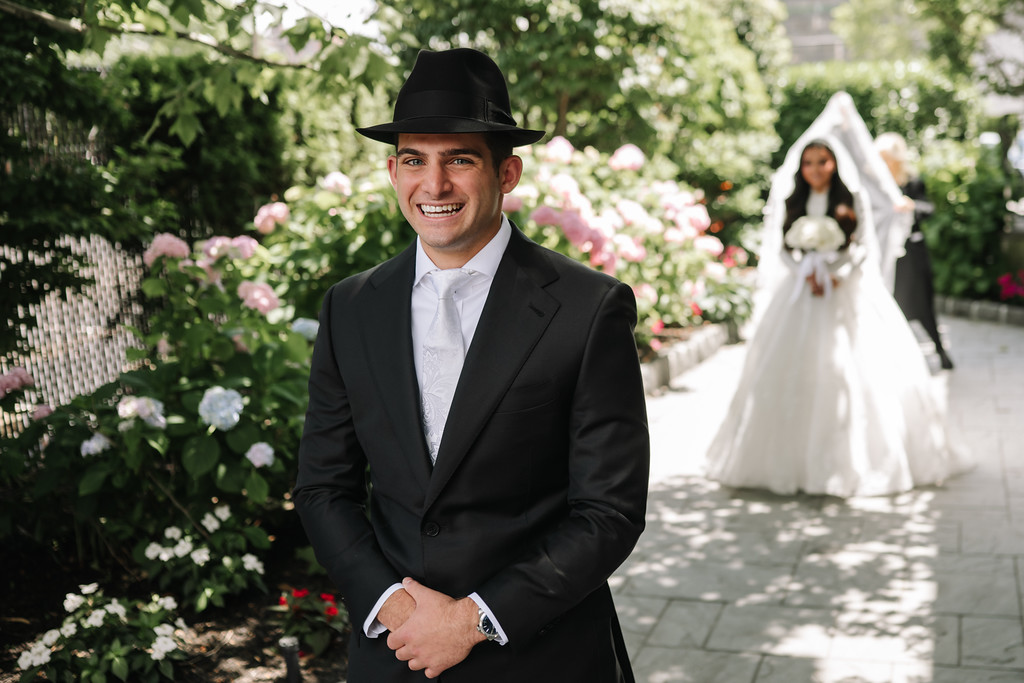 Outdoor wedding portrait of a smiling groom in a black hat and suit, with the bride in white in soft focus behind him.