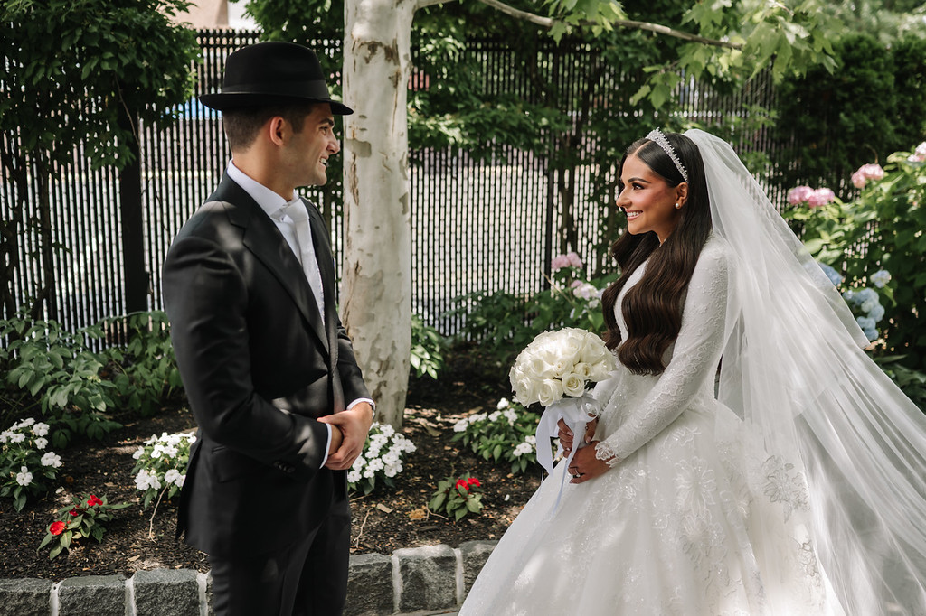 Jewish wedding couple standing outdoors during the badeken moment, groom in a black hat and suit facing the bride who wears a white veil and holds a bouquet of white flowers.