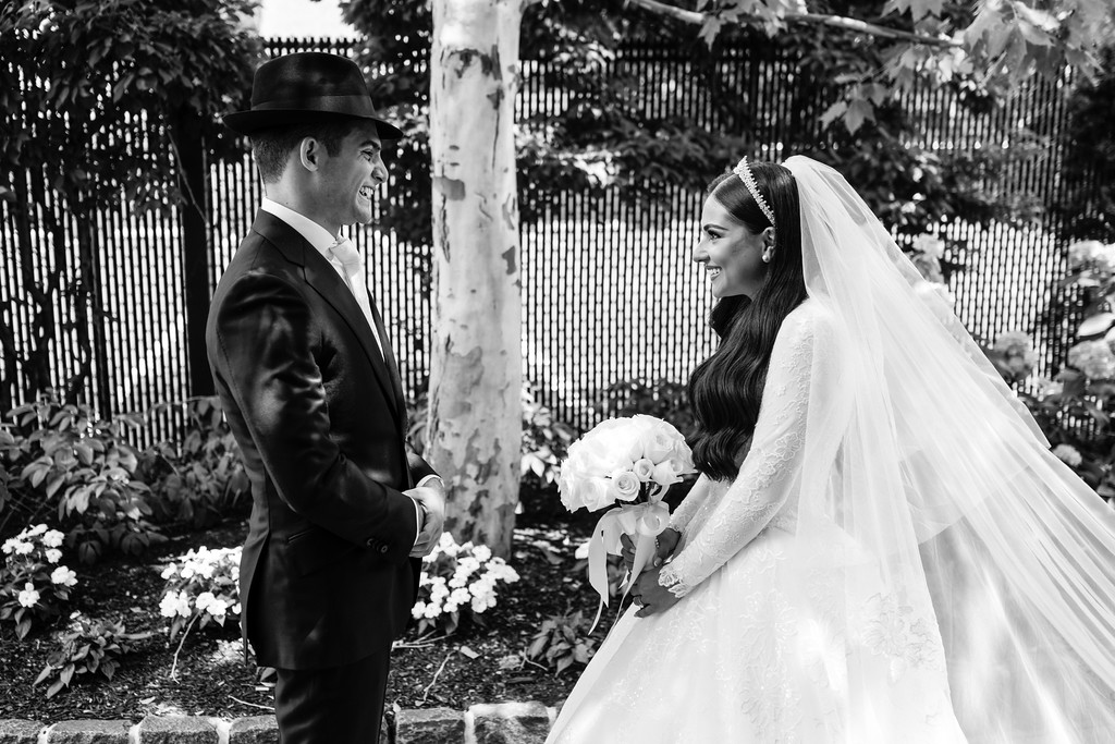 Black-and-white chuppah moment: groom in a black hat and suit stands facing the bride in a white wedding dress and veil as they smile at each other, bouquet in her hands.
