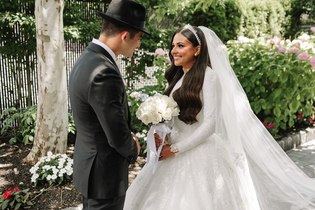 The groom in a black hat looks at the bride smiling in her white wedding dress and veil while holding a bouquet in a garden setting.