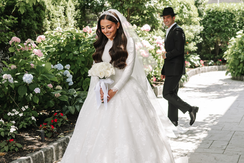 Smiling bride in a white gown and veil holding her bouquet while walking outdoors along a garden path; groom follows in the background.