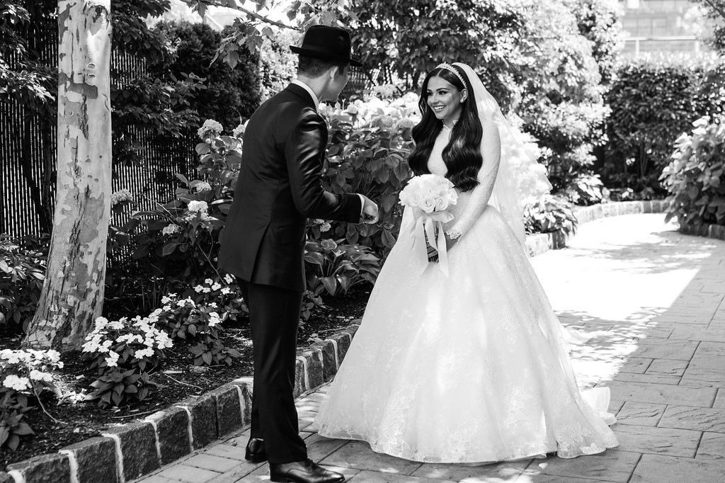 Black-and-white wedding photo of the groom and bride during the badeken, standing outdoors near flowers and greenery with the bride smiling under a veil.
